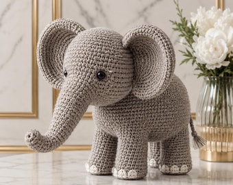 May include: A gray crocheted elephant toy featuring large rounded ears, a long trunk, and black bead eyes. The elephant has four sturdy legs with white crocheted details on the feet and a small tail with a tassel at the end of the body.