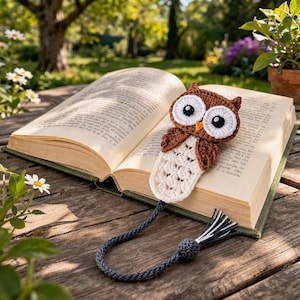 May include: A crocheted owl bookmark with a brown body, white eyes, and a cream-colored bottom section, resting on an open book. The bookmark has a dark gray cord with a tassel. The book is open on a wooden surface, with a blurred background of greenery.
