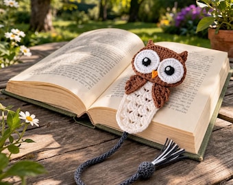 May include: A crocheted owl bookmark with a brown body, white eyes, and a cream-colored bottom section, resting on an open book. The bookmark has a dark gray cord with a tassel. The book is open on a wooden surface, with a blurred background of greenery.