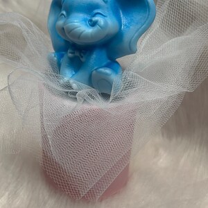 Può includere: Una candela a forma di elefante azzurro con un volto sorridente e un piccolo papillon. La candela è posta su una base rosa, avvolta in tulle bianco, e si trova su una superficie bianca e soffice.