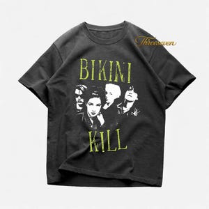 May include: Dark gray t-shirt featuring a band portrait and the words "BIKINI KILL" in a distressed, lime green font. The band members are depicted in a black and white graphic. The shirt has a crew neck and short sleeves.