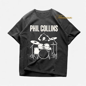 Può includere: T-shirt grigio scuro con grafica bianca di Phil Collins che suona la batteria, con il nome "PHIL COLLINS" in bianco sopra. La maglietta ha girocollo e maniche corte.