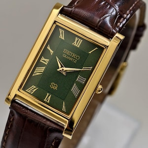 May include: A gold-toned Seiko Quartz watch with a rectangular green face and gold Roman numerals. The watch has a brown leather band. The watch face displays the Seiko Quartz logo. The watch is a classic, elegant design.