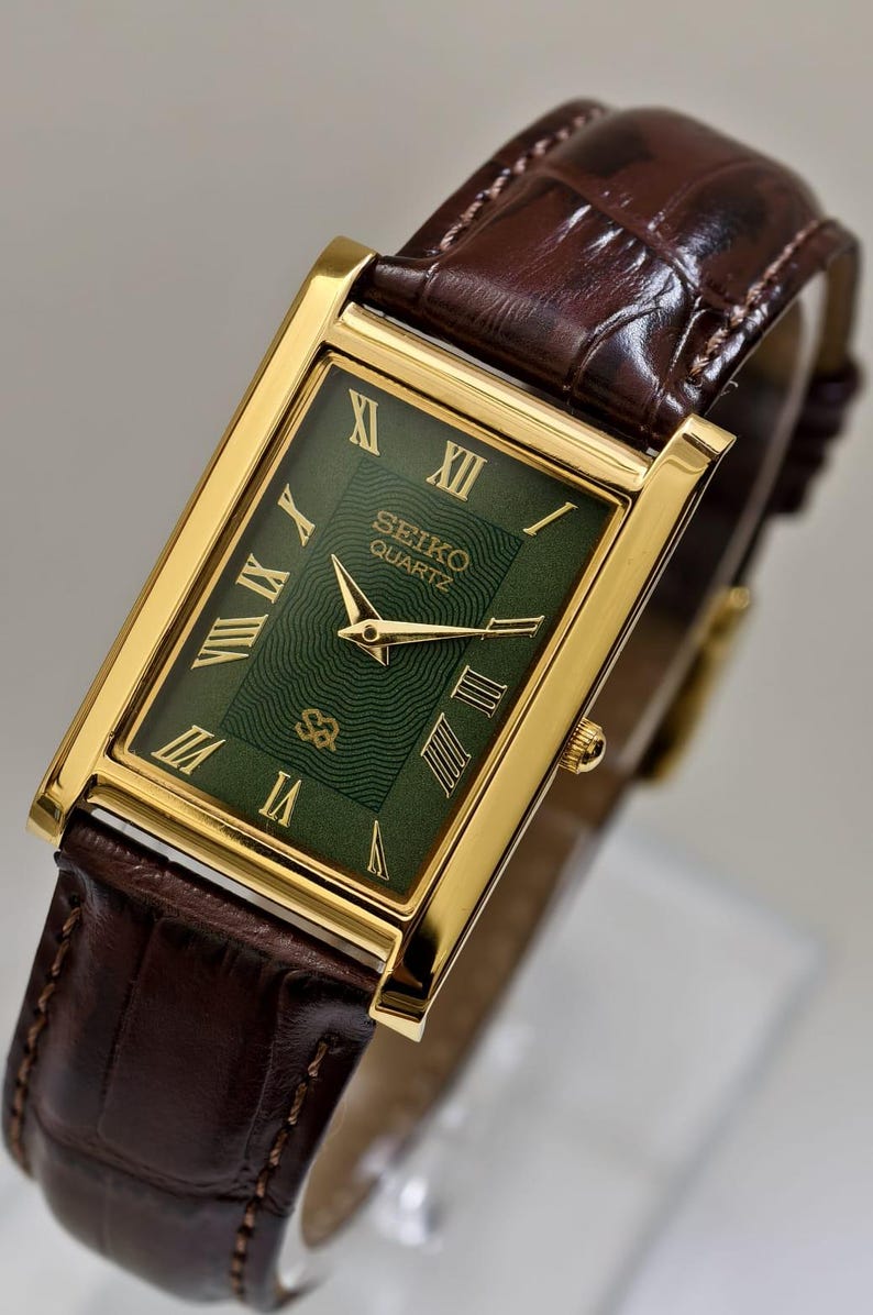 May include: A gold-toned Seiko Quartz watch with a rectangular green face and gold Roman numerals. The watch has a brown leather band. The watch face displays the Seiko Quartz logo. The watch is a classic, elegant design.