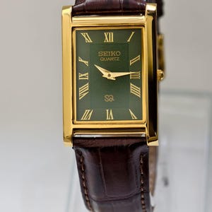 May include: A rectangular Seiko Quartz watch with a gold-tone case and a dark green dial. The watch features gold hands and Roman numeral hour markers. The brown leather band has a crocodile pattern.