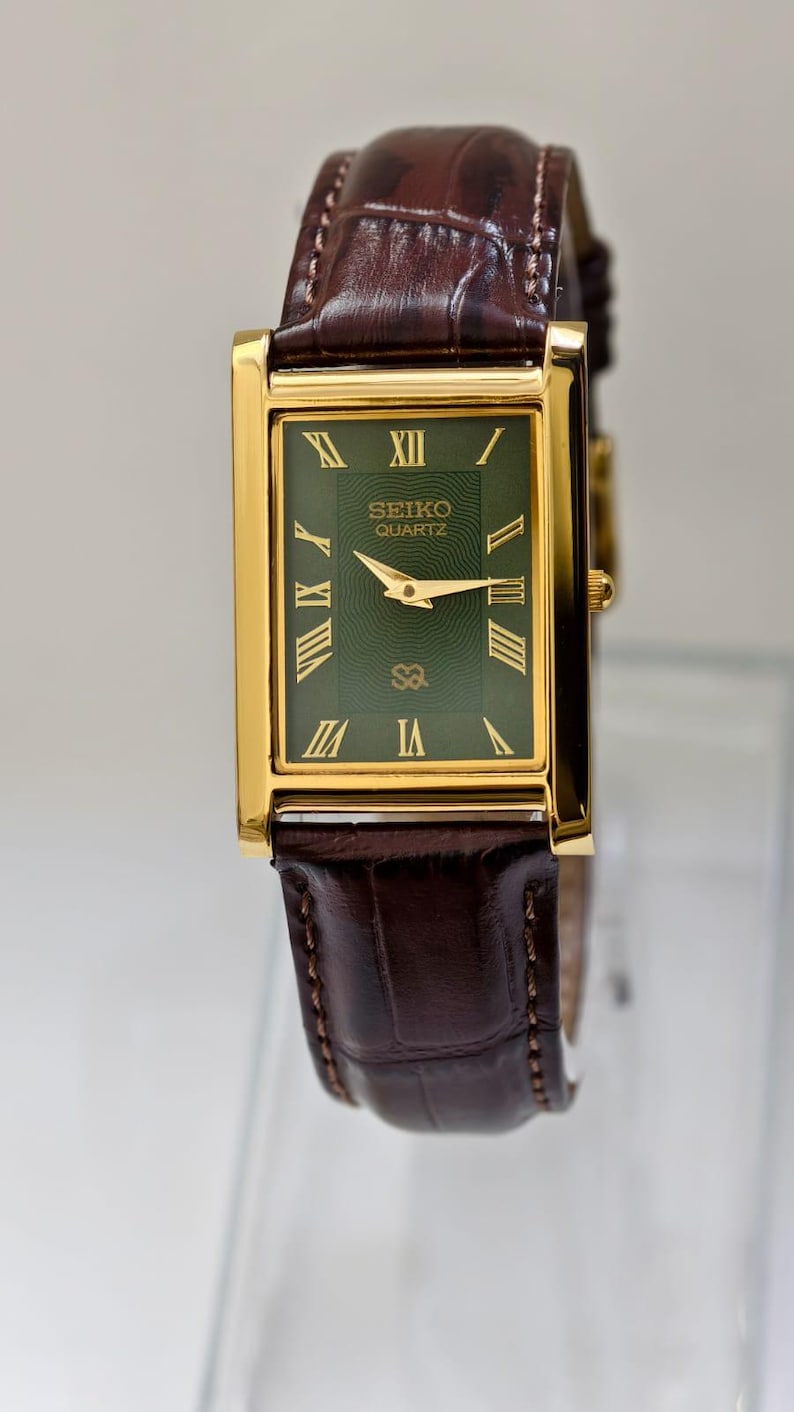 May include: A rectangular Seiko Quartz watch with a gold-tone case and a dark green dial. The watch features gold hands and Roman numeral hour markers. The brown leather band has a crocodile pattern.