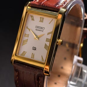May include: A gold-toned Seiko Quartz watch with a rectangular face and a brown leather strap. The watch face is white with gold Roman numerals and hands. The watch band is a rich brown color.