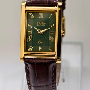 May include: A rectangular gold-toned Seiko Quartz watch with a green face and Roman numeral hour markers. The watch has gold hands and a brown leather band. The watch face reads "SEIKO QUARTZ".