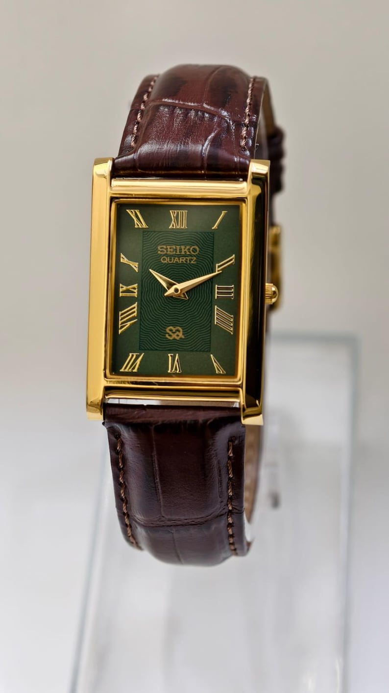 May include: A rectangular gold-toned Seiko Quartz watch with a green face and Roman numeral hour markers. The watch has gold hands and a brown leather band. The watch face reads "SEIKO QUARTZ".