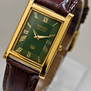May include: A gold-toned Seiko Quartz watch with a rectangular green face and gold Roman numerals. The watch has a brown leather band with a crocodile pattern. The watch face displays the Seiko Quartz logo.
