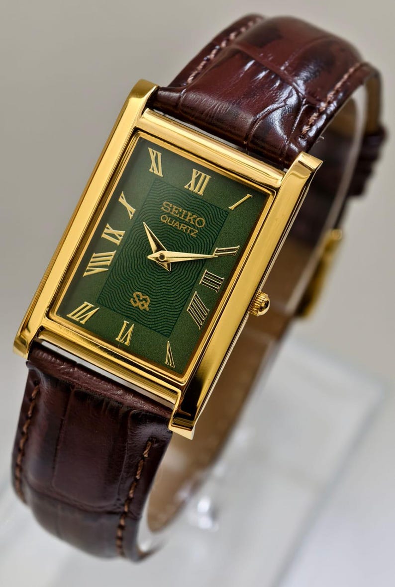 May include: A gold-toned Seiko Quartz watch with a rectangular green face and gold Roman numerals. The watch has a brown leather band with a crocodile pattern. The watch face displays the Seiko Quartz logo.