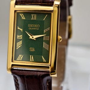 May include: A gold-toned Seiko Quartz watch with a rectangular green face and Roman numeral hour markers. The watch has gold hands and a brown leather band with a crocodile pattern. The watch is a classic timepiece.