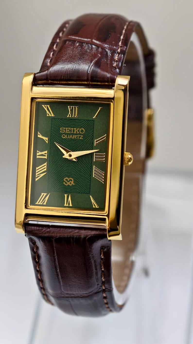 May include: A gold-toned Seiko Quartz watch with a rectangular green face and Roman numeral hour markers. The watch has gold hands and a brown leather band with a crocodile pattern. The watch is a classic timepiece.