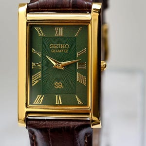 May include: A gold-toned rectangular Seiko Quartz watch with a green face and Roman numerals. The watch has gold hands and a brown leather band. The watch face has the Seiko Quartz logo.