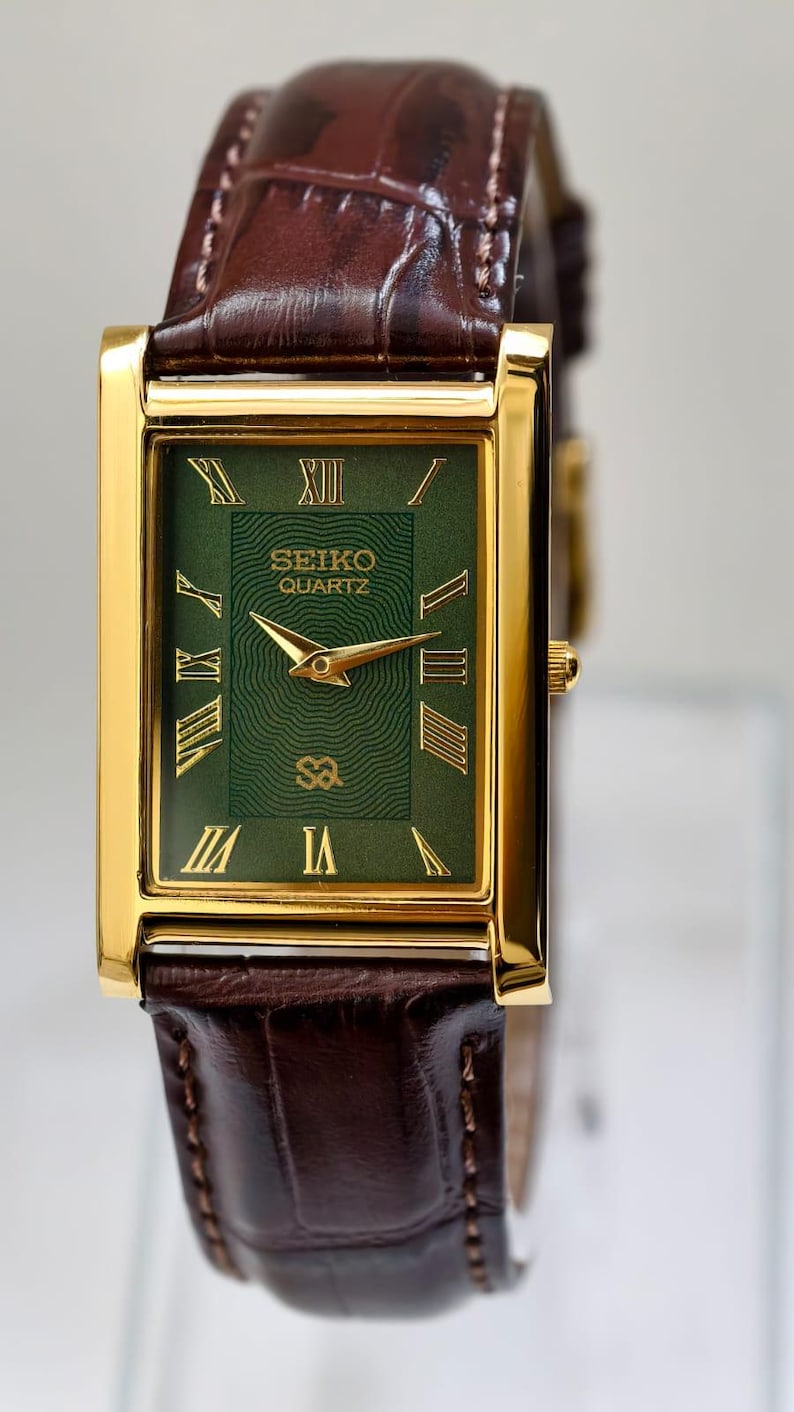 May include: A gold-toned rectangular Seiko Quartz watch with a green face and Roman numerals. The watch has gold hands and a brown leather band. The watch face has the Seiko Quartz logo.
