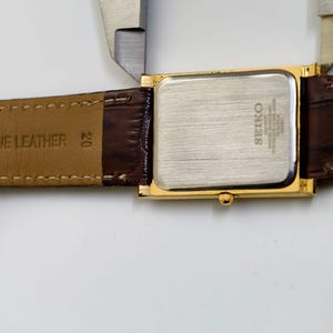 May include: A gold-toned Seiko quartz watch with a rectangular face and brown leather band. The watch is being measured with a silver caliper. The watch band is marked "GENUINE LEATHER".