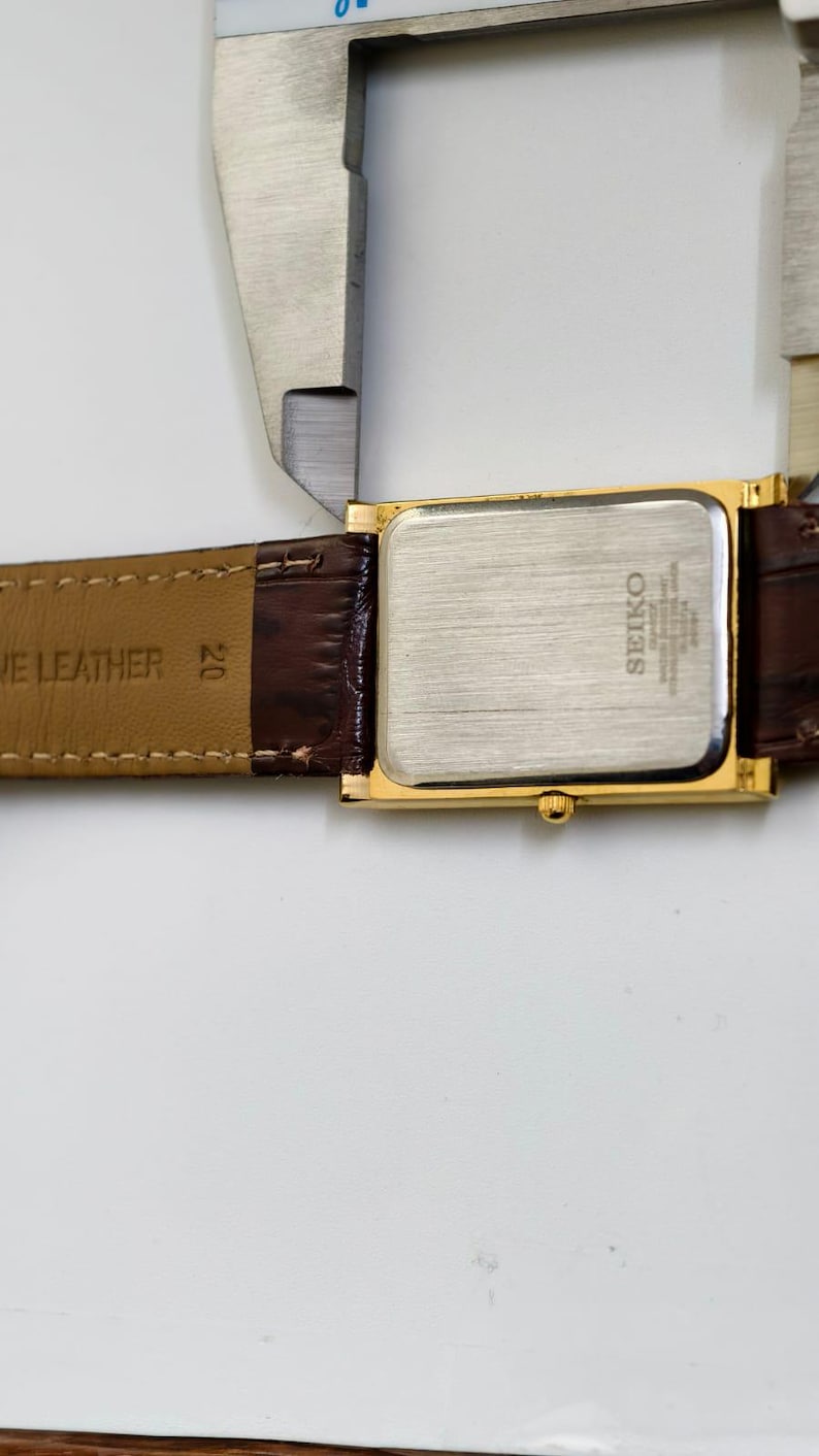 May include: A gold-toned Seiko quartz watch with a rectangular face and brown leather band. The watch is being measured with a silver caliper. The watch band is marked "GENUINE LEATHER".
