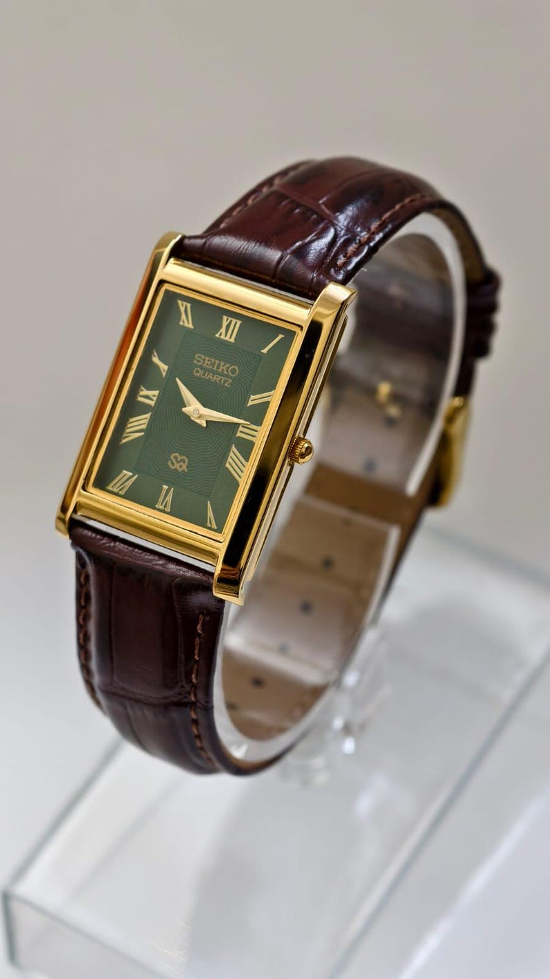 May include: A rectangular Seiko Quartz watch with a gold-tone case and a dark green dial. The watch features gold hands and Roman numeral hour markers. It has a brown leather strap with a crocodile pattern.