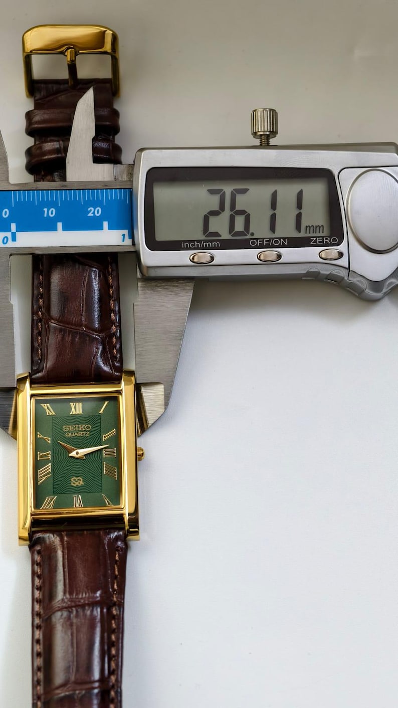 May include: A gold-toned Seiko Quartz watch with a rectangular green face and Roman numeral hour markers. The watch has a brown leather band and is measured at 26.11 mm. The watch is displayed with a digital caliper.