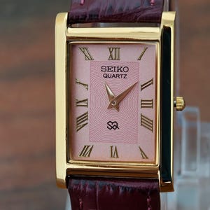 Puede incluir: Reloj Seiko Quartz dorado, rectangular, con esfera rosa y números romanos. Correa de cuero burdeos. La esfera muestra el logotipo de Seiko Quartz y un pequeño emblema.