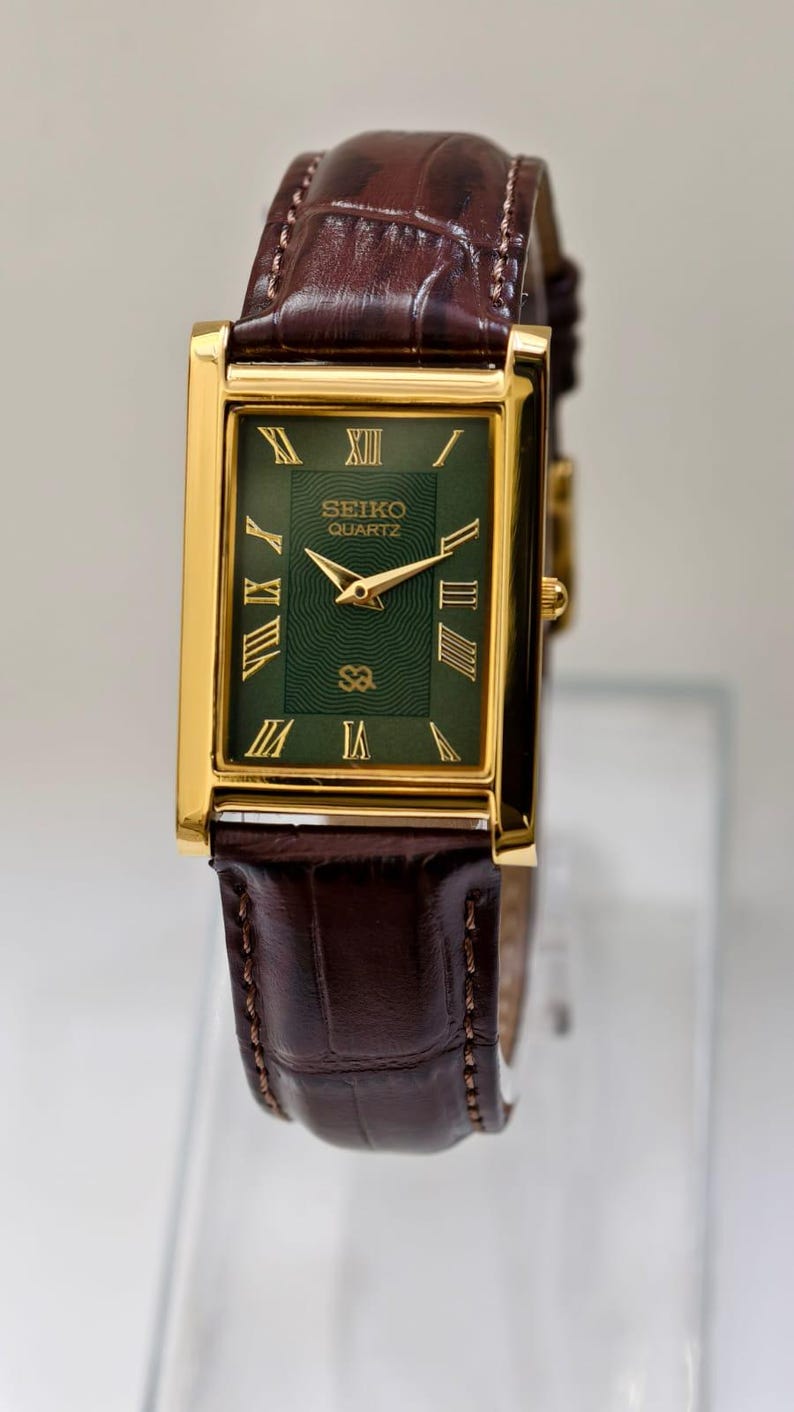 May include: A rectangular gold-toned Seiko Quartz watch with a green face and Roman numeral hour markers. The watch has a brown leather band. The watch face displays the Seiko Quartz logo.
