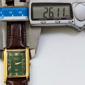 May include: A gold-toned Seiko Quartz watch with a rectangular green face and Roman numeral hour markers. The watch has a brown leather band and is measured at 26.11 mm. The watch is displayed with a digital caliper.