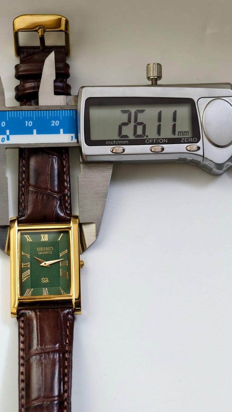 May include: A gold-toned Seiko Quartz watch with a rectangular green face and Roman numeral hour markers. The watch has a brown leather band and is measured at 26.11 mm. The watch is displayed with a digital caliper.