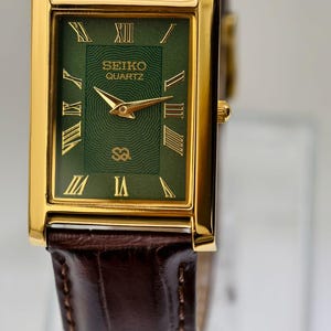 May include: A rectangular gold-toned Seiko Quartz watch with a green face and Roman numeral hour markers. The watch has gold hands and a brown leather band. The watch face displays the Seiko Quartz logo.