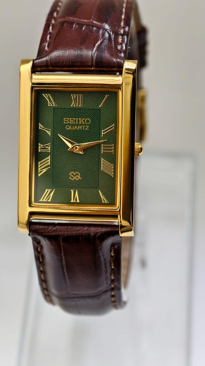 May include: A rectangular gold-toned Seiko Quartz watch with a green face and Roman numeral hour markers. The watch has gold hands and a brown leather band. The watch face displays the Seiko Quartz logo.