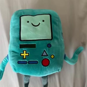 May include: A teal plush toy shaped like a retro video game console character. The toy has a smiling face on a screen, with arms, legs, and various colored buttons. The plush is soft and cuddly, perfect for fans of the animated series.