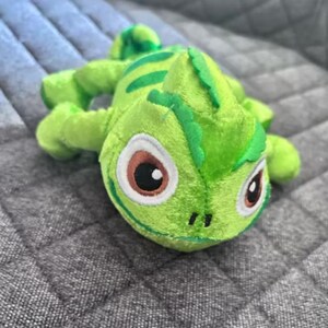 Puede incluir: Un peluche verde con forma de camaleón con detalles. El juguete presenta grandes ojos con detalles marrones y blancos, y una espalda texturizada. El camaleón está sobre una superficie gris acolchada.