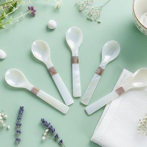May include: Five white mother-of-pearl spoons with a decorative band, arranged on a light green surface. The spoons are near a white bowl, white napkin, and sprigs of flowers. The spoons are approximately 6 inches long.