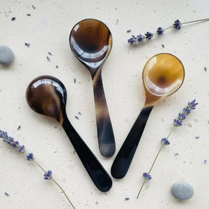 Może przedstawiać: Trzy ręcznie robione łyżki z ciemnobrązowymi i beżowymi wzorami, ułożone na jasnej powierzchni. Gałązki lawendy i małe kamienie są rozrzucone wokół łyżek. Łyżki mają błyszczące wykończenie i ciemny uchwyt.