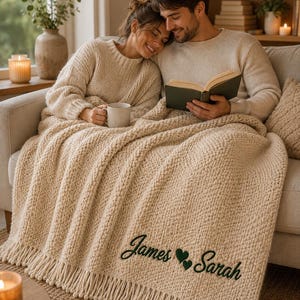 May include: A chunky knit beige throw blanket featuring a fringe edge and dark green embroidered text that reads James followed by a heart icon and Sarah. The blanket is draped over a sofa, showcasing a textured, thick yarn construction.