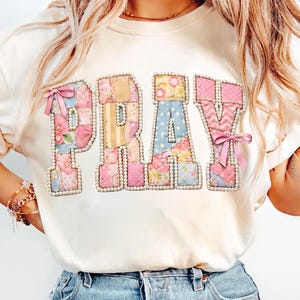 May include: A cream-colored t-shirt featuring the word PRAY in large block letters. Each letter is filled with a patchwork of floral and polka dot patterns in pink, blue, and yellow, outlined with white faux pearls and accented with pink bows.