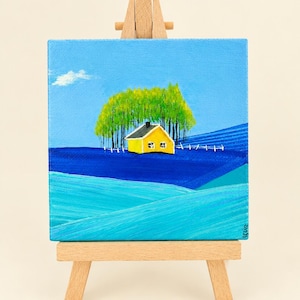Può includere: Un piccolo dipinto su tela quadrata su un cavalletto di legno raffigura una casa giallo brillante con il tetto nero immersa tra alti alberi verdi. Il paesaggio presenta colline ondulate in tonalità blu e turchese sotto un cielo azzurro.
