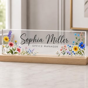 Könnte beinhalten: Ein rechteckiges Schreibtisch-Namensschild aus klarem Acryl mit dem aufgedruckten Text Sophia Miller und OFFICE MANAGER in Schwarz. Das Design ist beidseitig mit bunten Wildblumenillustrationen verziert und steht auf einem hellen Holzsockel.