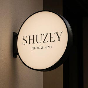 Könnte beinhalten: Ein rundes, beleuchtetes Schild mit schwarzem Rahmen ist an einer beigen Wand befestigt. Das Schild zeigt den Namen "SHUZEY" in einer fetten, schwarzen Schrift über "moda evi" in kleinerer Schrift. Das Schild ist mit einem weichen, warmen Licht beleuchtet.