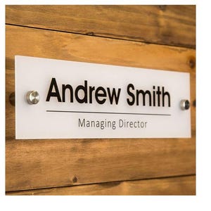 以下が含まれることがあります： Andrew SmithとManaging Directorという黒い文字が書かれた長方形の白いアクリル製サインプレート。名前と肩書きの間に水平線があり、左右に2つの円形の銀色の金属製スタンドオフで固定されています。