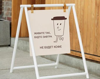 May include: A-frame sign with a white metal frame and a beige sign. The sign features a cartoon coffee cup with a smiling face and the Russian text "Live like tomorrow won't have coffee."