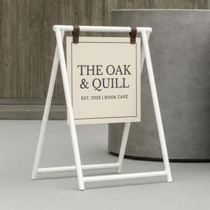 Może przedstawiać: Biały metalowy szyld w kształcie litery A z prostokątnym szyldem w kolorze kremowym. Na szyldzie widnieje napis "THE OAK & QUILL EST. 2023 | BOOK CAFÉ" czarnym tekstem. Szyld jest utrzymywany na miejscu przez brązowe skórzane paski.