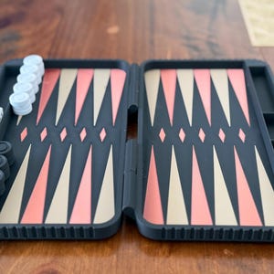 May include: A portable backgammon set featuring a dark grey folding case with a playing surface of alternating coral and beige triangles. The set includes white and dark grey circular checkers and two white dice with black pips arranged on the board.