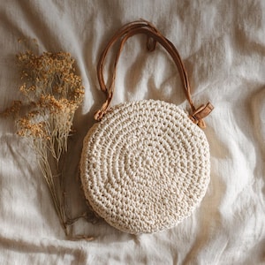 Puede incluir: Un bolso redondo de crochet color crema con asas de cuero marrón. El bolso es circular y tiene una superficie texturizada. Flores secas están al lado del bolso sobre una tela color crema.