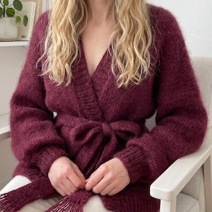 Könnte beinhalten: Burgunderfarbener Cardigan mit langen Ärmeln, Wickeldesign und passendem Bindegürtel mit Schleife. Der Cardigan ist aus einem dicken, strukturierten Strickstoff gefertigt. Die Ärmel sind lang und der Cardigan hat einen V-Ausschnitt.