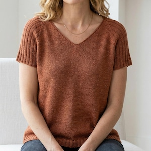 May include: A rust-colored short-sleeved sweater with a V-neck. The sleeves have a ribbed design, and the body of the sweater has a textured knit. A gold necklace is visible. The sweater is worn by a person sitting down.