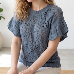 Può includere: Un top blu lavorato a maglia con maniche corte e arruffate. Il top presenta un motivo a foglie dettagliato sul corpo e sulle maniche. Lo scollo tondo e l'orlo a coste aggiungono un tocco classico a questo capo fatto a mano. Il top è di un morbido colore blu.