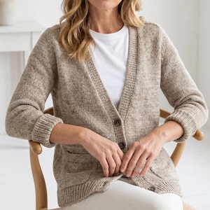 May include: A beige knit cardigan with a V-neck, button closure, and two front pockets. The cardigan has ribbed cuffs and a textured knit pattern. The person is wearing a white t-shirt and white pants. The cardigan is a neutral color.