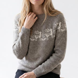 Peut inclure: Un pull gris avec un col rond et des poignets côtelés. Le pull présente un motif blanc de rennes sur la poitrine. Le modèle porte le pull, la main près du visage.
