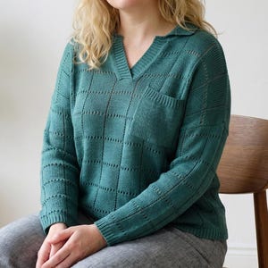 May include: Teal green knit sweater with a V-neck collar and a small pocket on the chest. The sweater has a grid pattern design. The person is wearing gray pants and sitting on a wooden chair.