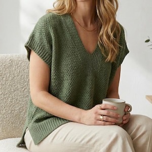 May include: Olive green knitted sleeveless top with a V-neck. The top features a textured pattern and ribbed trim. The person is wearing a gold necklace and rings, and holding a beige mug. The person is wearing beige trousers.